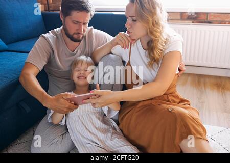 happy family with daughter watch funny video on mobile phone, girl is laughing, sit on the floor in living room