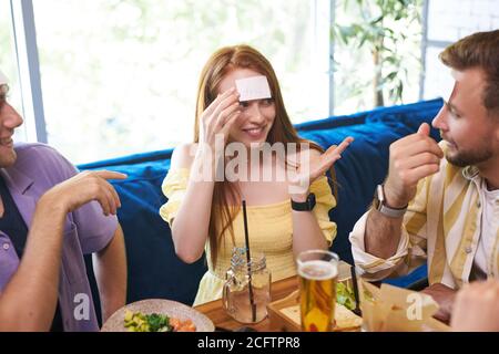 little sticker on the forehead. friends having fun in the restaurant, caucasian young people playing a hedbanz game, laugh Stock Photo