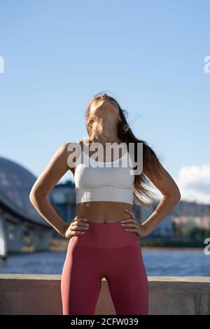 Fit young athletic woman stretching body on embankment. Fitness female in pink leggings doing warm up workout for neck muscles outdoor. Stock Photo