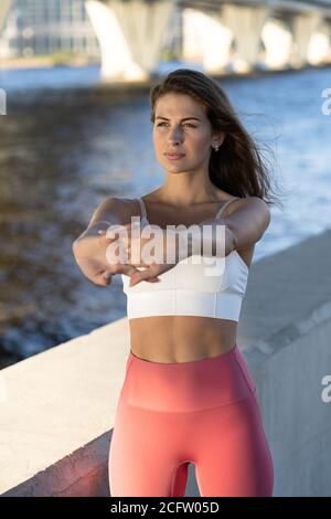 Fit young athletic woman stretching arms and body on embankment. Fitness female in pink leggings doing warmup workout outdoor. Stock Photo