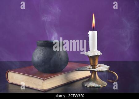 Candle in candlestick burning on a black desk and witch smoky pot on an old book on a purple background. Halloween concept Stock Photo