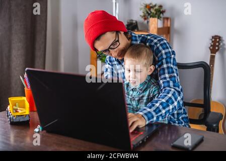 Young modern man father is working on a laptop, and his little son is sitting on his lap. Concept of family and remote work from home Stock Photo
