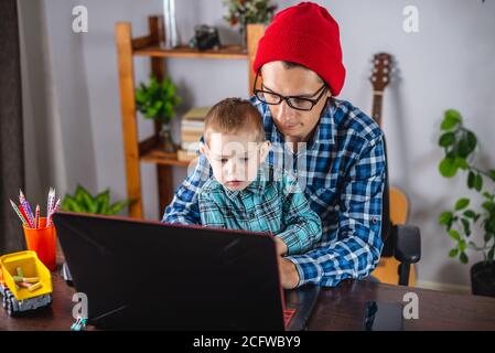 Young modern man father is working on a laptop, and his little son is sitting on his lap. Concept of family and remote work from home Stock Photo
