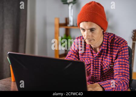 Modern young man in a red checked shirt and hat is working on a laptop. Concept of distance communication and remote job. Stock Photo