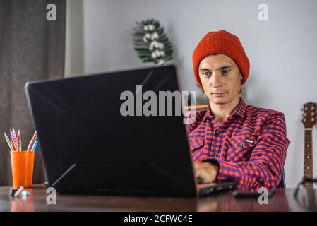 Modern young man in a red checked shirt and hat is working on a laptop. Concept of distance communication and remote job. Stock Photo
