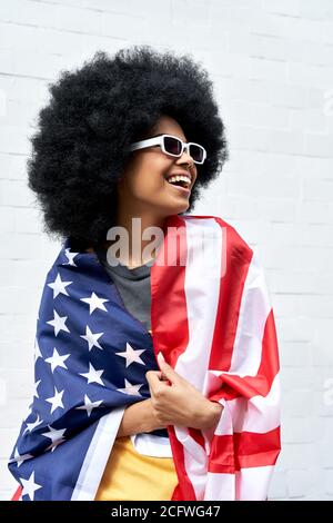Happy young African woman wrapped in USA flag standing against white wall. Stock Photo