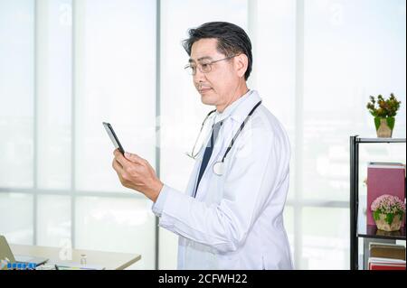 Asian adult  male doctor wearing formal white coat using a smartphone for working service. Healthcare specialist people. Stock Photo