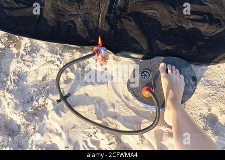 A woman with air foot pump pumps an inflatable mattress or air bed at sandy beach. Foot inflates air mattress with foot pump on sand. Stock Photo