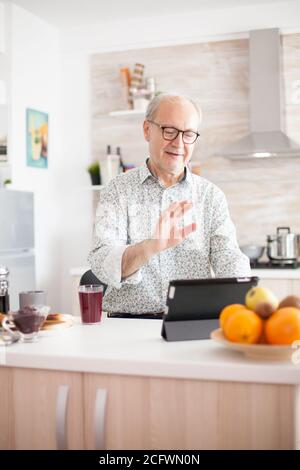 Happy senior man during breakfast in kitchen on a video conference with his family. Elderly person using internet online chat technology video webcam making a video call connection camera communication conference call Stock Photo