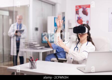 Doctor wearing high tech virtual reality goggles in hospital office and white coat with stethoscope. Senior medical writing patient diagnostic on clipboard and nurse is holding sick man x-ray image. Stock Photo