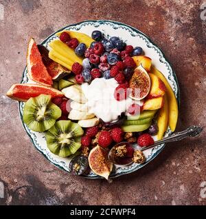 Assorted sliced fruits on vintage plate. Mango, kiwi fruits, figs, grape, bananas, raspberries and blueberries with yoghurt and granola. Stock Photo