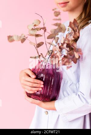 Girl in a blue linen dress posing on a pink background. Girl holding a purple glass vase with dry plant branches in her hands Stock Photo