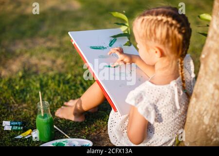 Cute little caucasian kid girl drawing or painting with colored paints in park. Kindergarten children education, back to school after coronavirus Stock Photo