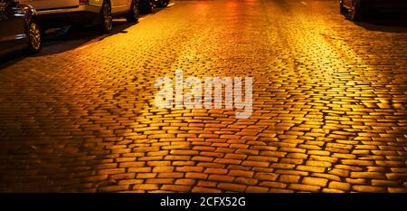Paving stones with reflection of yellow light at night. Night city abstract background and texture. Cobblestone road at night illumination. Soft focus Stock Photo