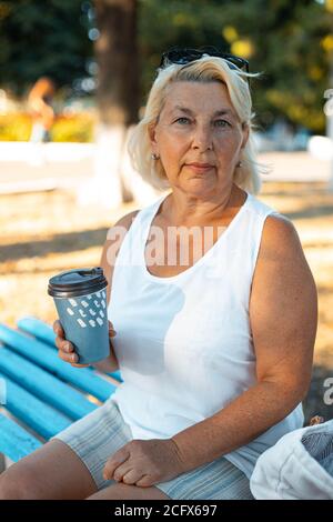 Adult blonde smiling woman with cup of takeaway coffee sitting on ood bench in green park on summer day, Stock Photo