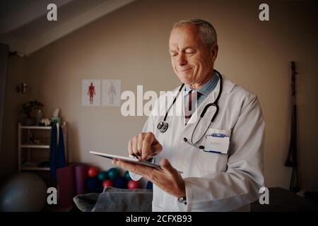 Standing senior doctor with stethoscope around neck holding digital tablet in hospital - happy Doctor using technology  Stock Photo