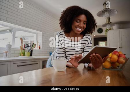 African American Woman standing in kitchen leaning on counter using digital tablet looking at online recipes  Stock Photo