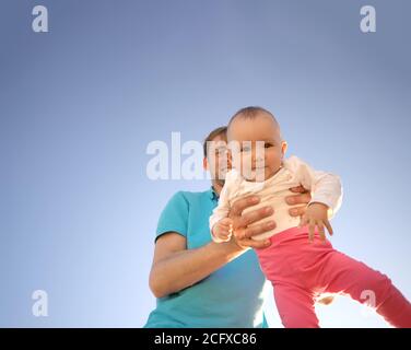 Father holds a small child in his arms on a background of a summer blue sky. Cute baby is smiling. Happy family outdoors. Stock Photo