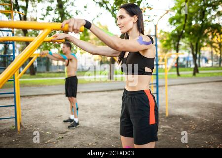 Side view of caucasian athletes couple with kinesiology elastic taping on bodies, brunette woman and man training using bars at sports ground. Concept Stock Photo