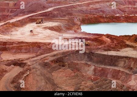 Aluminium ore quarry and blue lake in bauxite mine. Yellow excavators and quarry trucks. White road. Open cast (open-cut) mining. Quarry lake. Stock Photo