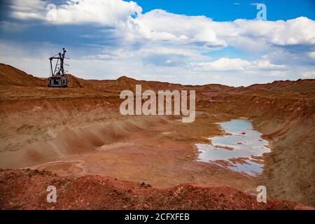 Walking dragline excavator in bauxite clay quarry. Heaps of minerals and small lake. Aluminium ore mining. On blue sky with clouds. Stock Photo