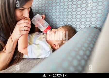 Mother gives baby bottle of milk on sofa as concept for love and care Stock Photo