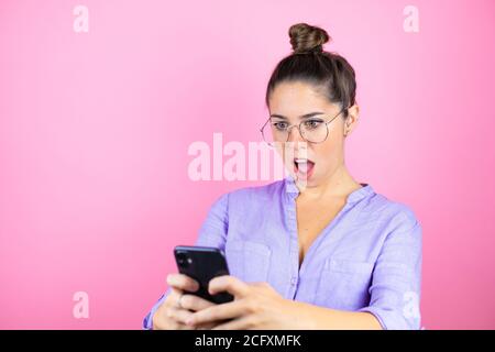 Young beautiful woman wearing glasses over isolated pink background chatting with her phone and surprised Stock Photo