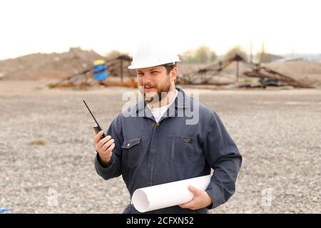 Portrait of caucasian foreman standing on construction site, talking by VHF walkie talkie and holding blueprints. Stock Photo