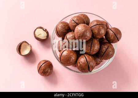 Raw macadamia nuts in a transparent glass bowl and scattered on pink solid background. Minimal composition. Stock Photo