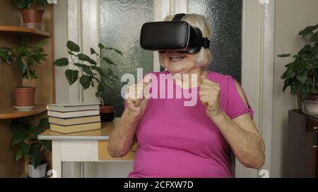 Amazed senior grandmother in virtual headset glasses watching amazing 3d video in 360 vr helmet at home. Future technology. Woman in VR goggles playing games, looking around, shows emotion of surprise Stock Photo
