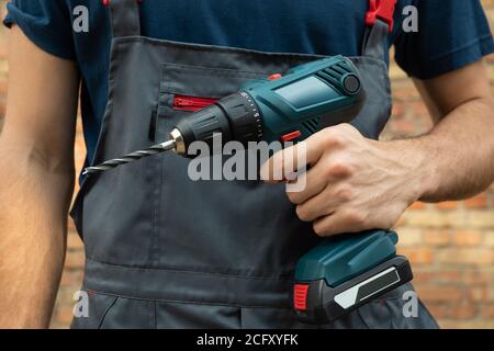 Man in overalls holds cordless drill on brick wall background Stock Photo