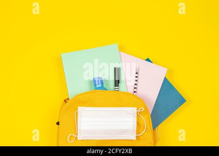 Education, learning, back to school during pandemic. Top view to backpack with school supplies, hand sanitizer, protective mask on yellow background Stock Photo