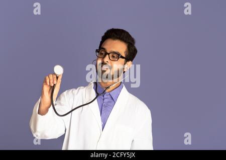 Young Indian doctor in lab coat using stethoscope on lilac background, free space Stock Photo