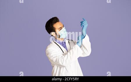 Protection against virus. Indian doctor in medical mask and lab coat putting on rubber gloves on lilac background Stock Photo