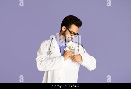 Corruption in healthcare concept. Indian doctor putting money into his pocket on lilac background Stock Photo