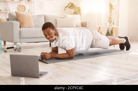African Guy Training At Laptop Doing Plank Exercise In Bedroom Stock Photo