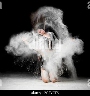 two beautiful young caucasian women in black bodysuits with a sports figure are dancing in a white cloud of flour on a black background, explosion and Stock Photo