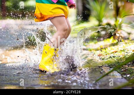 Kid playing in the rain in autumn park. Child jumping in muddy puddle on rainy fall day. Little girl in rain boots and rainbow jacket outdoors in heav Stock Photo