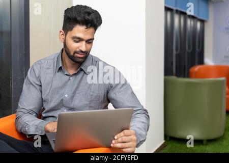 Portrait of young successful Indian business man sitting on couch and working on laptop, Corporate environment. Stock Photo