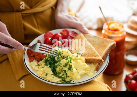 Eating scrambled eggs with baked beans and bread toast. Female hands holding plate with breakfast omelette. Eating food concept Stock Photo