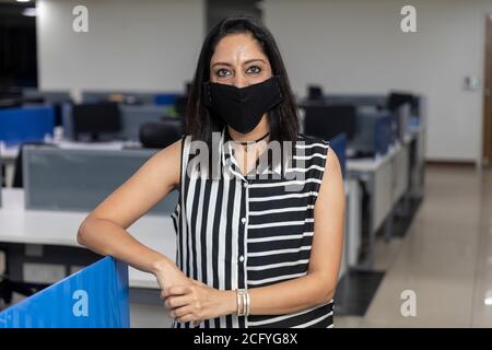 Portrait of successful Indian business woman wearing Covid protection mask and standing against office background, Corporate environment. Stock Photo