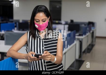 Portrait of successful Indian business woman wearing Covid protection mask and standing against office background, Corporate employee checking phone. Stock Photo
