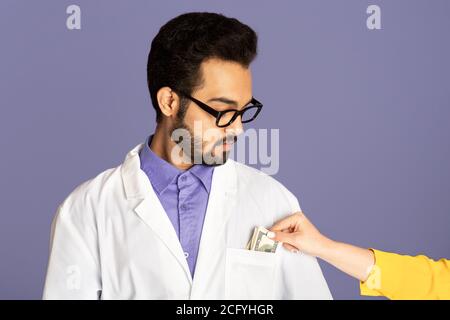 Corruption in medicine. Patient putting money into Indian doctor's pocket on lilac background Stock Photo