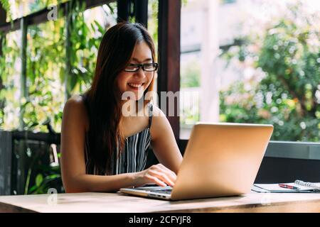 Positive Asian female student in glasses smiling and browsing laptop while sitting at table on terrace and studying on summer day Stock Photo