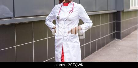 black young woman doctor, in a white coat, with a phonendoscope, holds a folder for medical papers in a rue, against the background of a hospital. Stock Photo