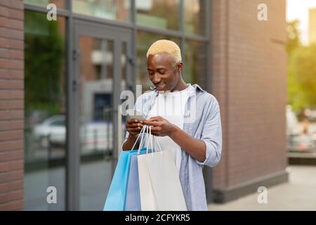 Smiling african american guy with packages with purchases in hands, typing on smartphone Stock Photo