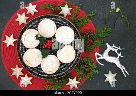 Delicious Christmas mince pies on a plate with winter berry holly, cedar cypress, mistletoe, silver star & reindeer decorations on grey grunge. Stock Photo