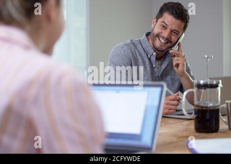 Business Couple Working From Home Sitting At Table During Pandemic Lockdown Using Mobile Phone Stock Photo