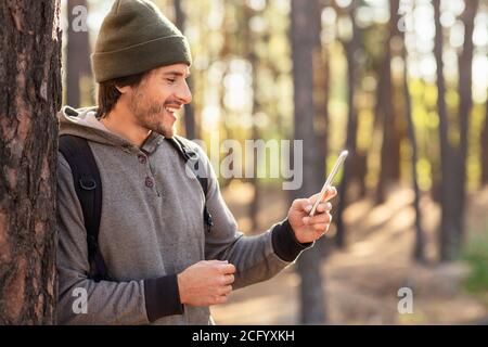 Positive guy leaning on tree in forest, using mobile phone Stock Photo