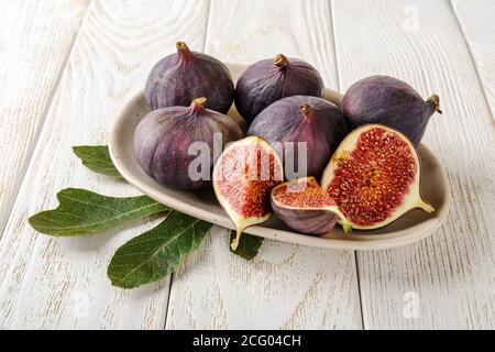 Whole and cutted fresh ripe fig fruits on a beige ceramic plate over a white wood table. Sweet fruits and berries. Vegetarian, raw food diet. Stock Photo